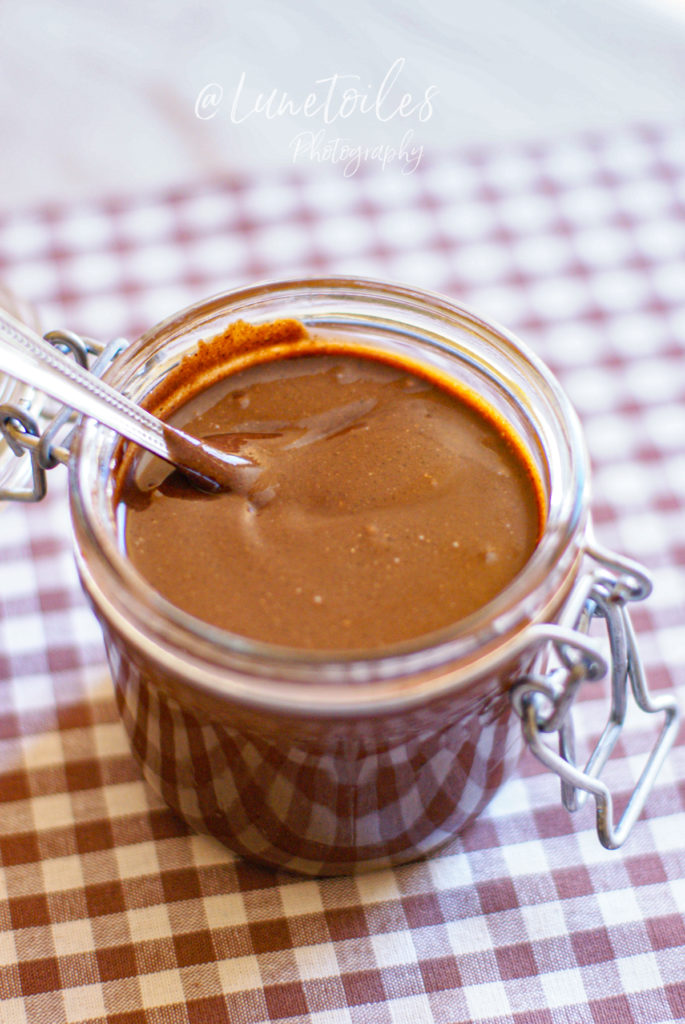 A glass jar filled with smooth, homemade chocolate hazelnut spread, with a spoon dipped inside, placed on a brown and white checkered tablecloth