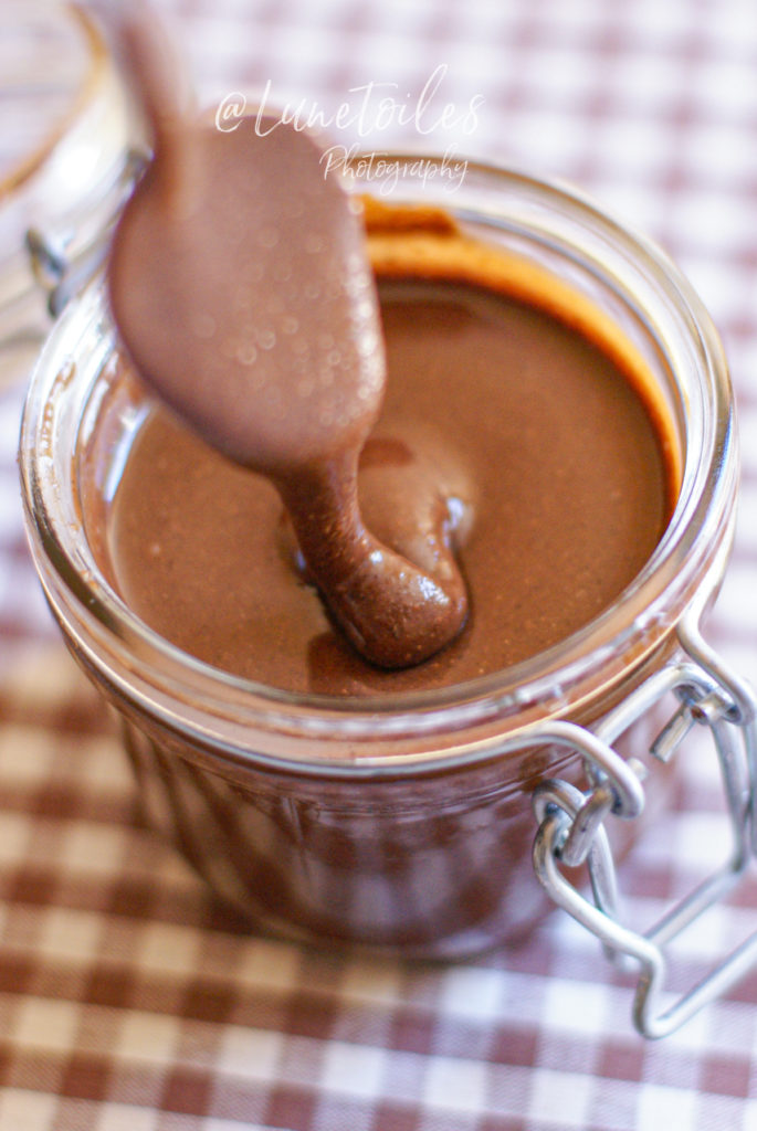 A close-up of a spoon lifting creamy, glossy homemade Nutella from a glass jar, set on a checkered cloth, highlighting the rich texture and indulgent chocolate-hazelnut color