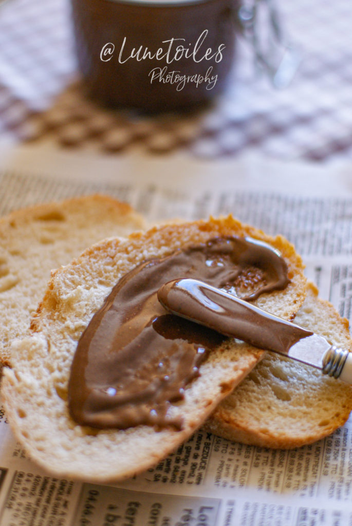 A slice of rustic bread topped with a generous layer of creamy homemade Nutella spread, with a butter knife resting on top, and a glass jar of the spread blurred in the background on a checkered tablecloth