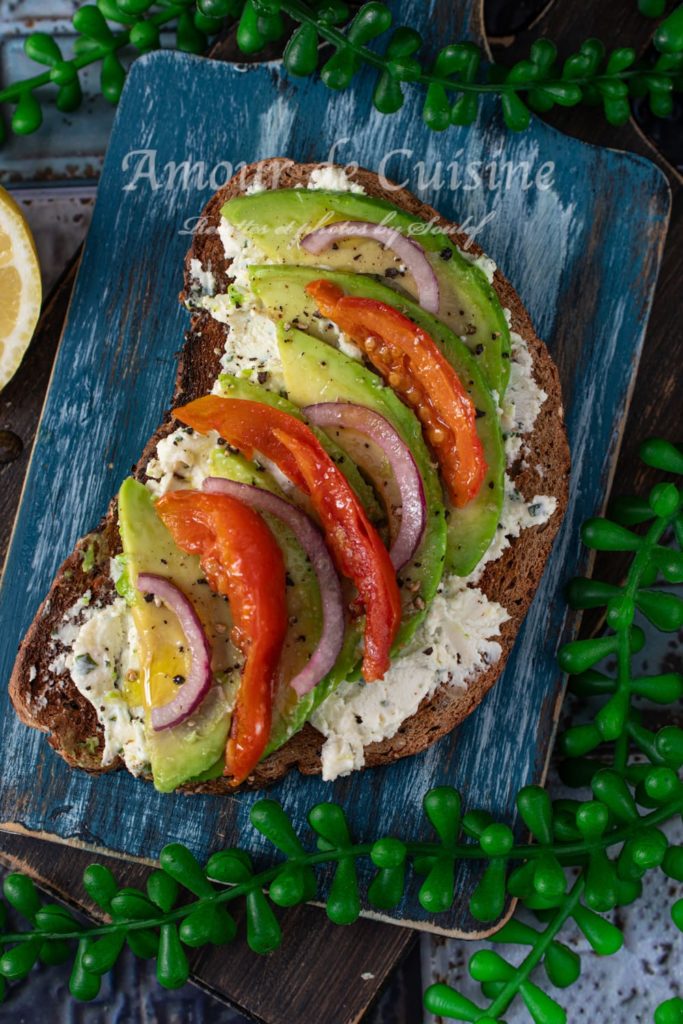 Avocado toast topped with creamy cheese spread, sliced avocado, roasted red tomatoes, and red onion on multigrain bread, served on a rustic blue wooden board with lemon and green decorative plants in the background