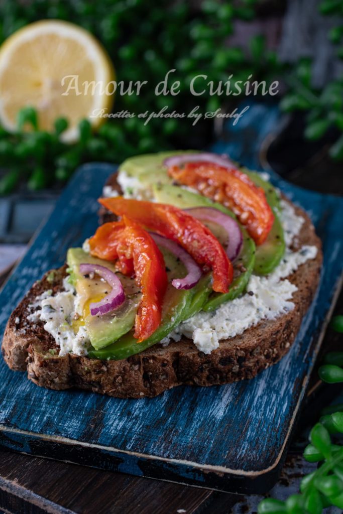 Avocado toast topped with sliced avocado, roasted red peppers, red onion, and a layer of creamy cheese on multigrain bread, served on a rustic blue wooden board with a lemon and greenery in the background