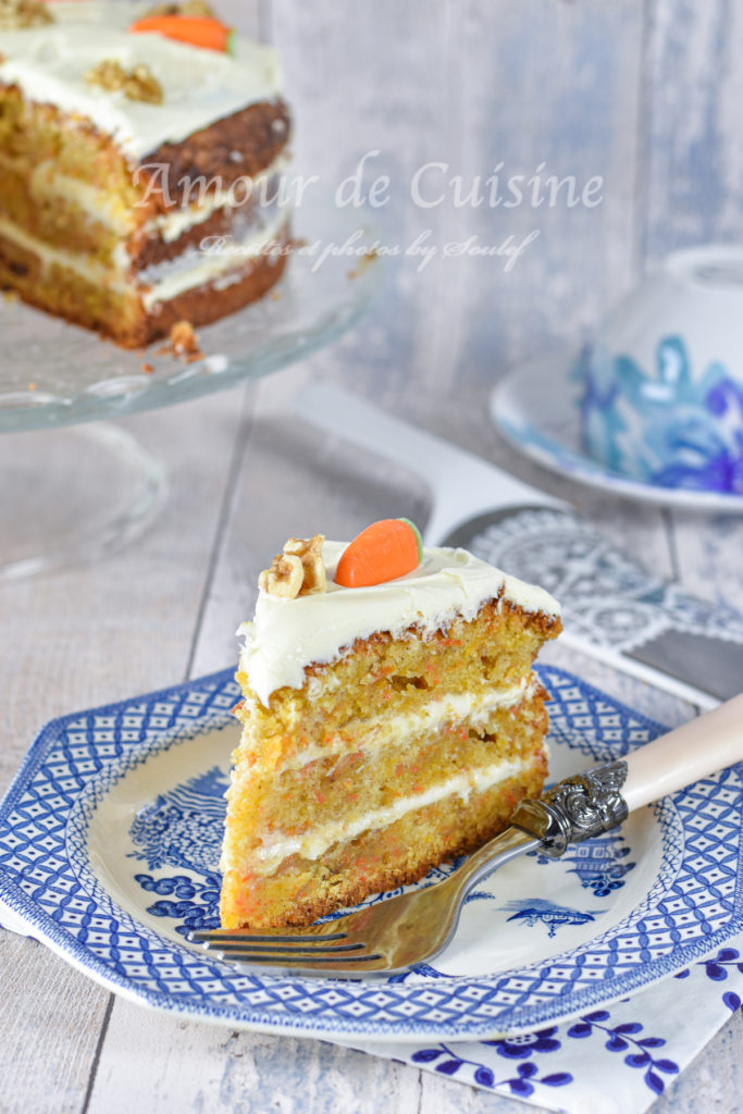 A slice of homemade carrot cake with cream cheese frosting and a decorative marzipan carrot on top, served on a blue and white patterned plate with a silver fork, with the rest of the cake visible in the background on a glass stand