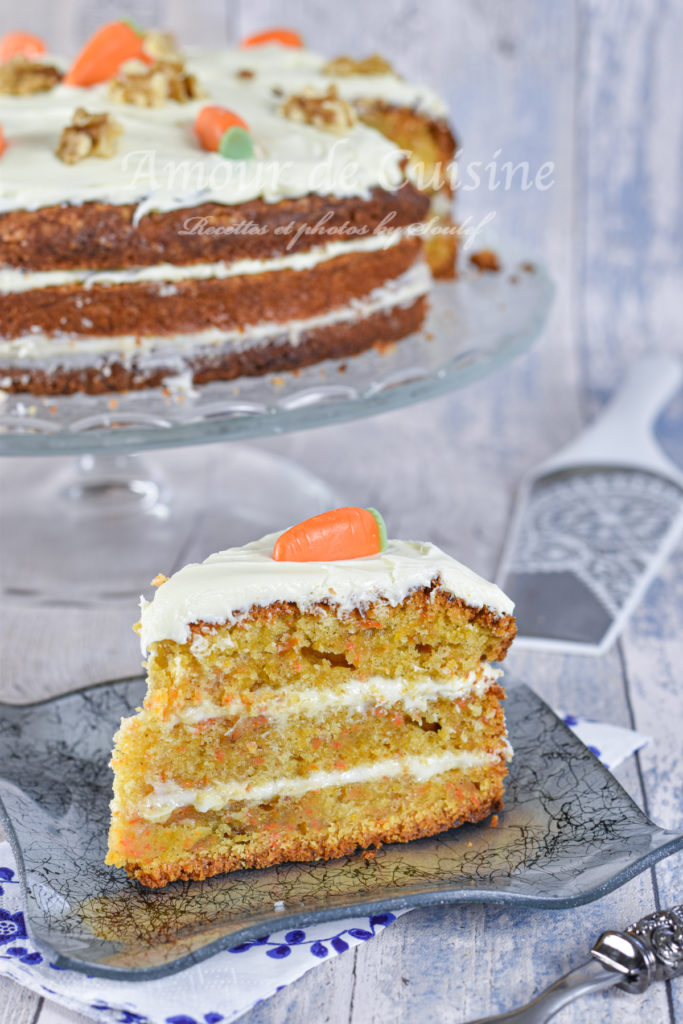 A slice of moist, three-layer carrot cake with cream cheese frosting and a small marzipan carrot on top, served on a decorative gray glass plate, with the full frosted cake in the background on a glass stand