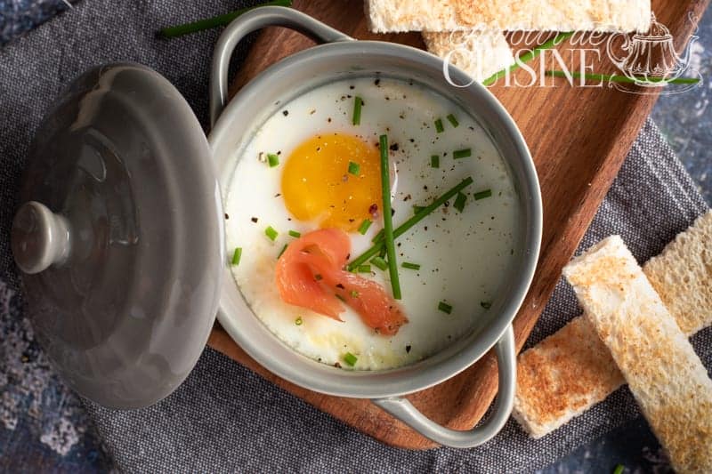 Top view of a baked egg en cocotte in a grey dish, garnished with smoked salmon, chopped chives, and black pepper, served with toasted breadsticks on a wooden tray