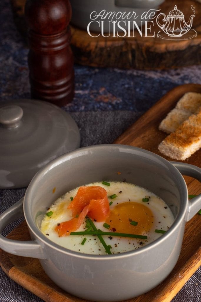 Eggs en cocotte served in a grey ceramic dish, topped with smoked salmon, chopped chives, and cracked pepper, placed on a wooden tray with toasted breadsticks and a pepper grinder in the background.