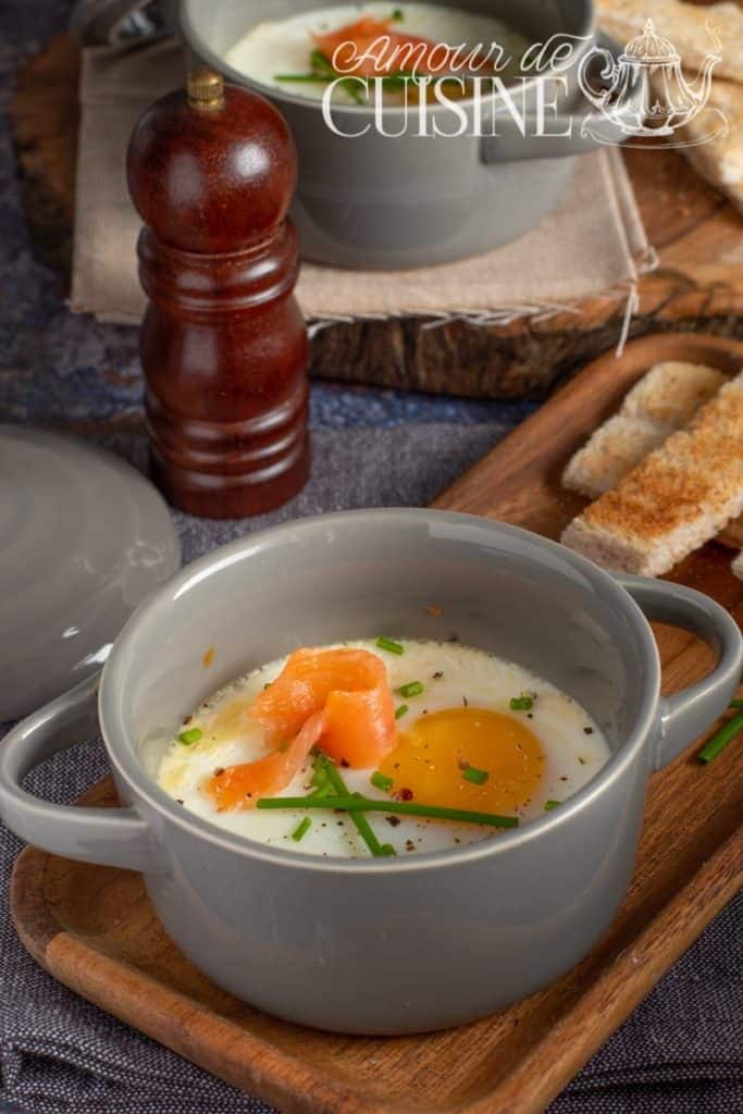 Baked eggs en cocotte served in a grey ramekin, topped with smoked salmon and chopped chives, placed on a wooden tray with toasted bread strips and a pepper grinder in the background.