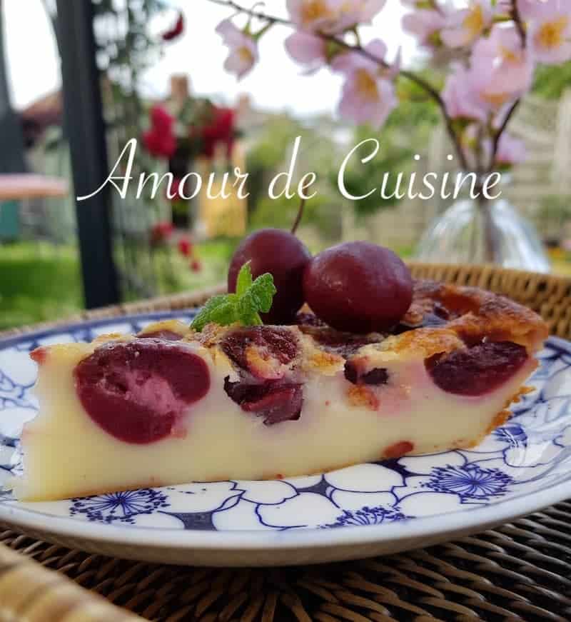 Slice of creamy cherry clafoutis on a floral-patterned plate, garnished with fresh cherries and mint, served outdoors with blooming flowers in the background