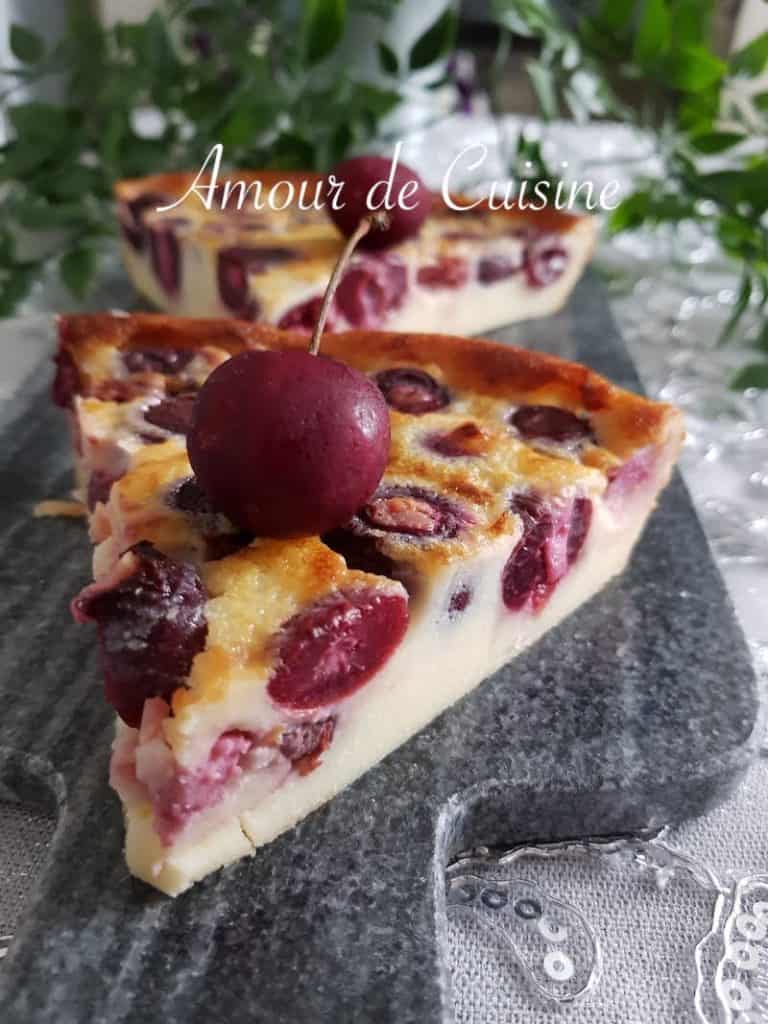 Slice of homemade cherry clafoutis with a golden top and juicy whole cherries, garnished with a fresh cherry on top, served on a stone serving board.