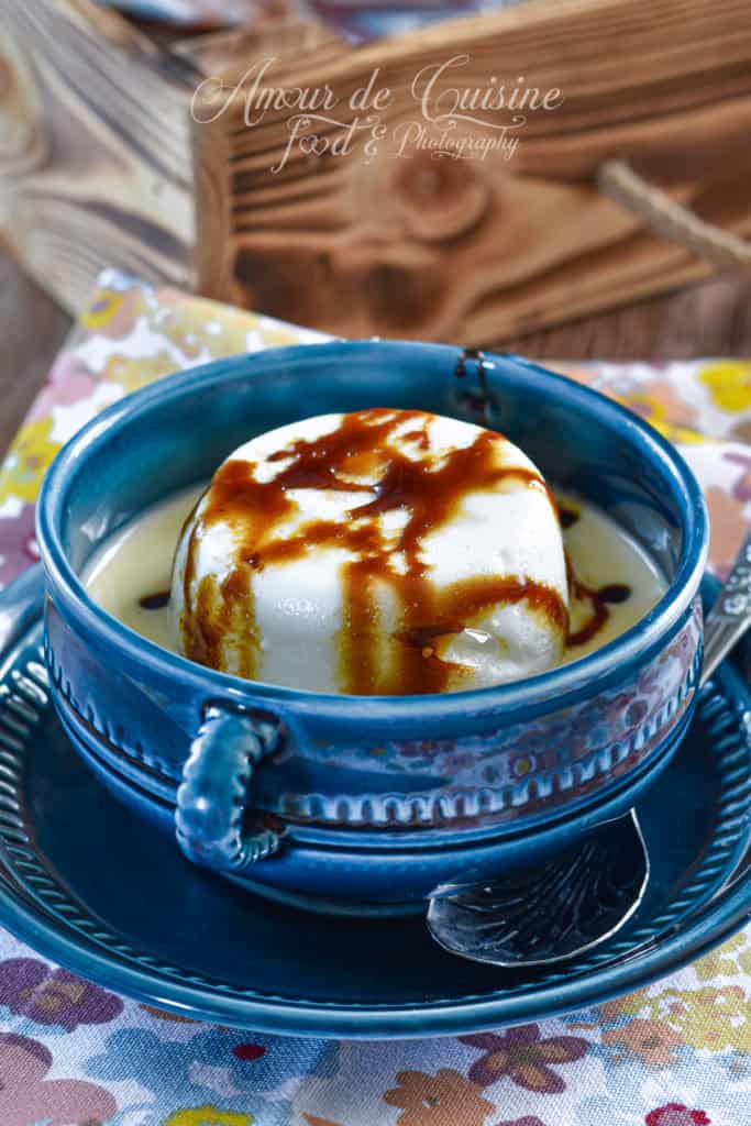 A traditional French floating island dessert (Île Flottante) served in a blue ceramic bowl, featuring a fluffy poached meringue topped with golden caramel sauce, floating on a bed of creamy vanilla custard. The bowl sits on a matching saucer with a spoon, atop a floral-patterned tablecloth