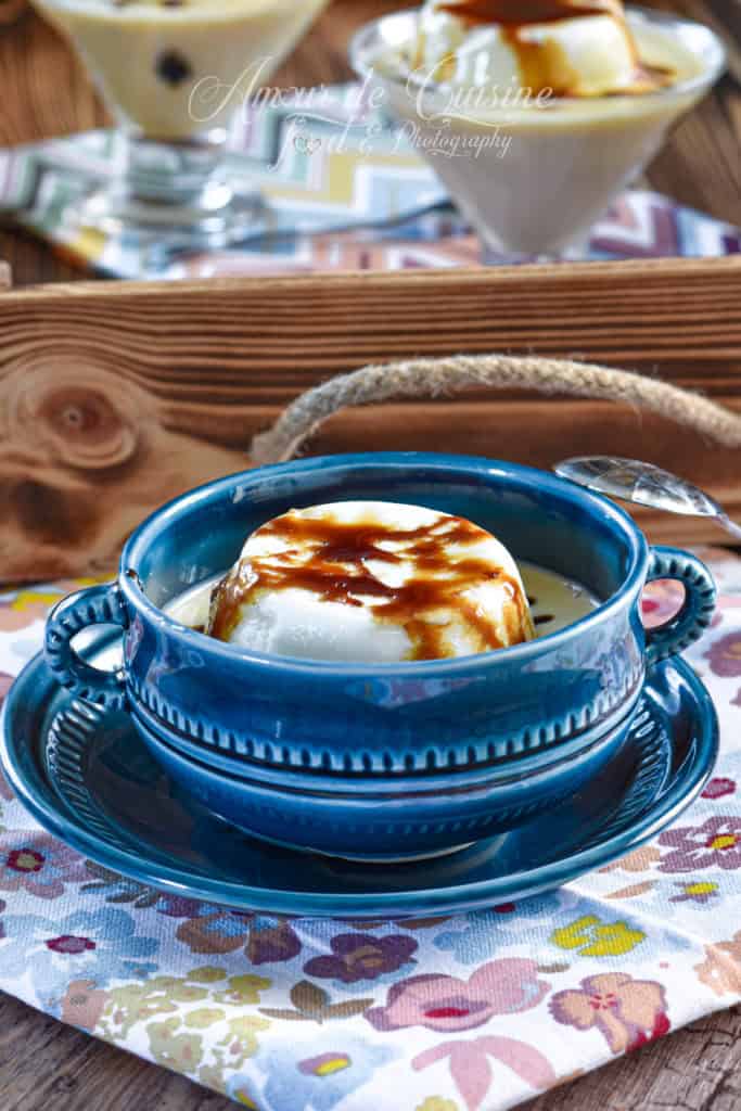 A rich and elegant serving of French Floating Island dessert (Île Flottante) presented in a deep blue ceramic bowl with matching saucer. The dish features a fluffy poached meringue drizzled with golden caramel, floating on creamy vanilla custard. In the background, additional servings are displayed in glass dessert cups, all placed on a floral-patterned tablecloth