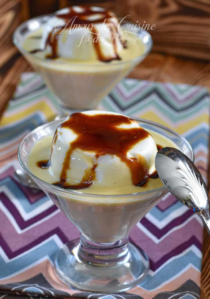 Two elegant glass dessert cups filled with French Floating Island (Île Flottante), featuring fluffy poached meringue floating on creamy vanilla custard and topped with rich caramel sauce. The desserts are served on a colourful chevron-patterned napkin, with a silver spoon resting beside the front cup