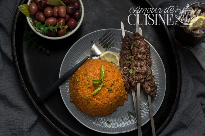 Grilled beef and lamb kefta skewers served with a dome of tomato bulgur, garnished with fresh herbs and lemon, accompanied by a bowl of olives and a drink with lemon slice