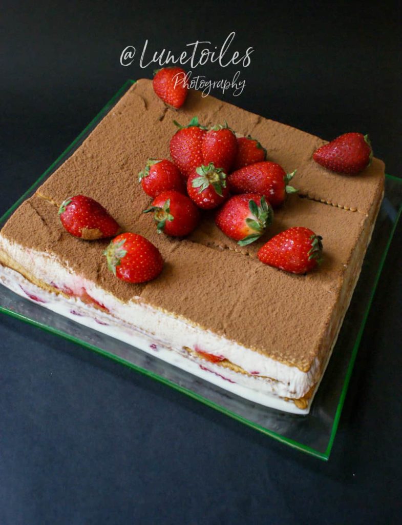 No-bake strawberry refrigerator cake layered with biscuits, cream, and strawberries, topped with cocoa powder and fresh whole strawberries, presented on a glass plate against a dark background.