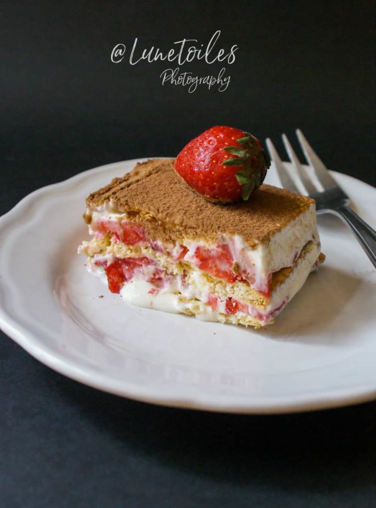 A slice of no-bake strawberry icebox cake served on a white plate, showing layers of biscuits, creamy filling, and fresh strawberries, topped with cocoa powder and a whole strawberry, with a fork placed beside it.