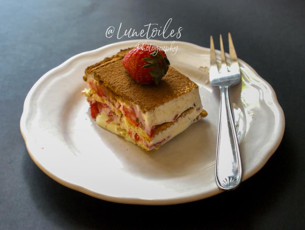 A slice of strawberry icebox cake on a white plate, showing layers of creamy filling, fresh strawberries, and biscuits, topped with a dusting of cocoa powder and a whole strawberry, served with a silver fork on the side