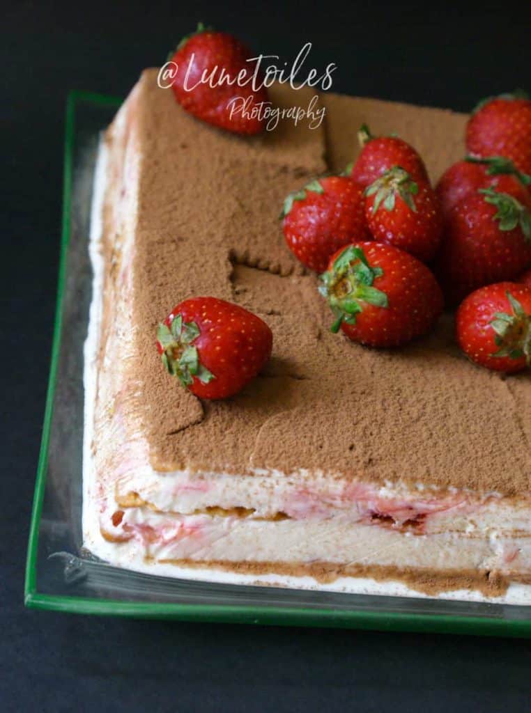 Close-up of a no-bake strawberry icebox cake in a glass dish, featuring visible creamy layers of whipped filling, biscuit, and strawberries, topped with a dusting of cocoa powder and garnished with fresh whole strawberries