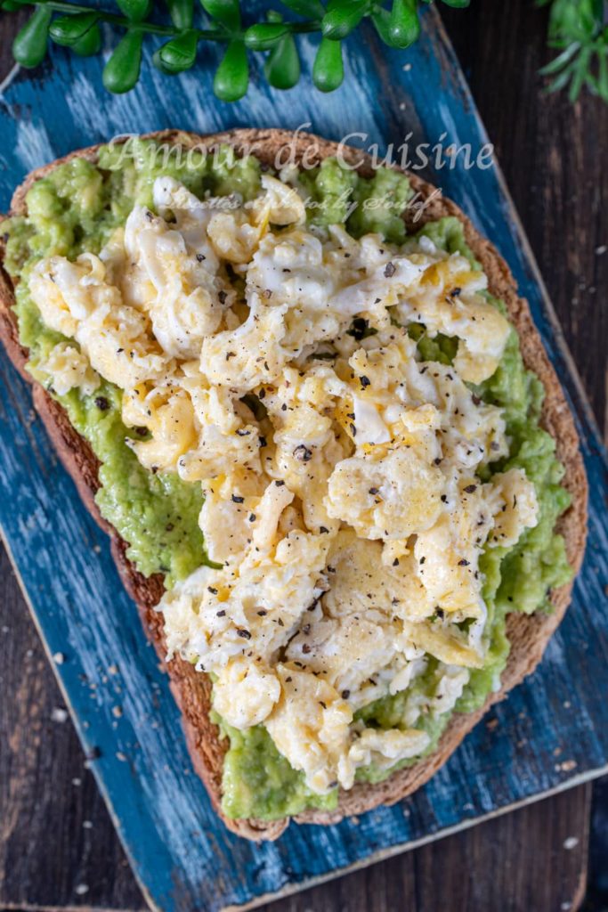 Close-up of scrambled egg avocado toast on whole grain bread, featuring creamy mashed avocado and fluffy scrambled eggs, seasoned with black pepper and served on a rustic blue wooden board with greenery in the background