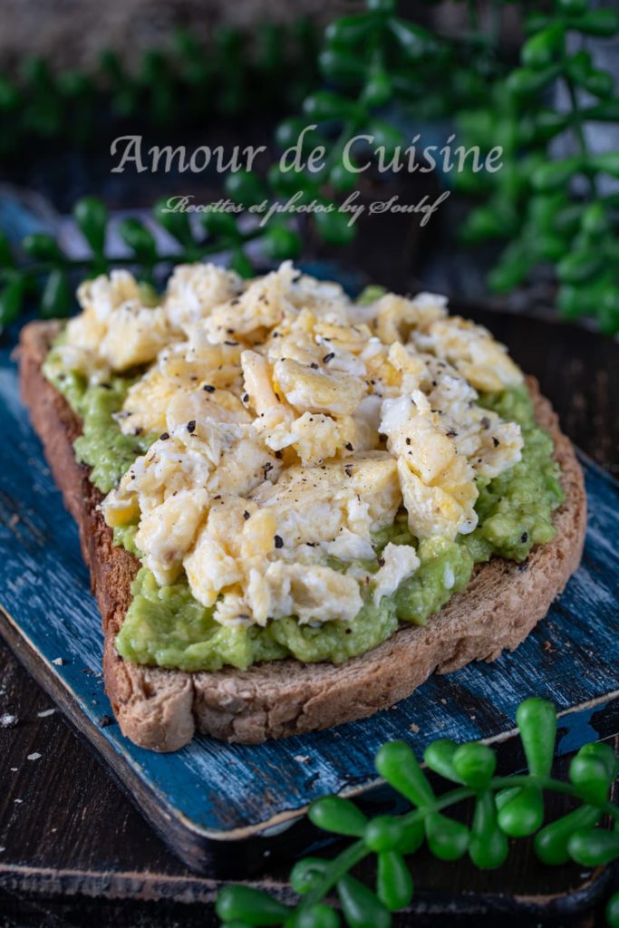 Avocado toast topped with creamy scrambled eggs and a sprinkle of black pepper on a slice of rustic whole grain bread, served on a distressed blue wooden board with green plant decor in the background