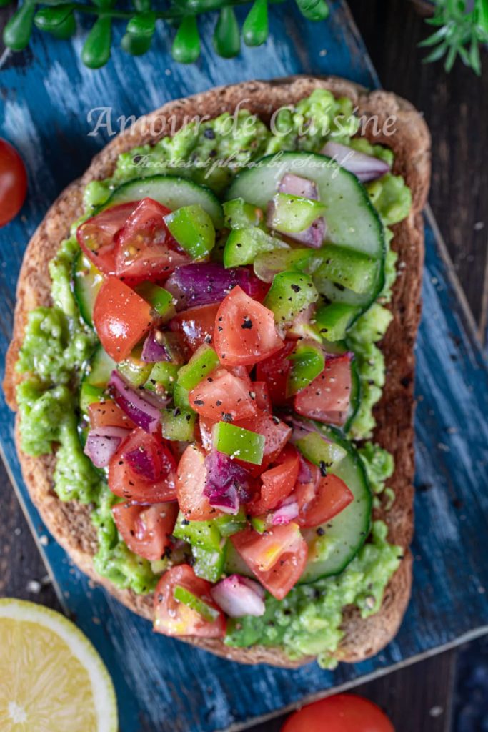 Avocado toast topped with mashed avocado, cucumber slices, diced cherry tomatoes, green bell pepper, and red onion, seasoned with black pepper, served on rustic bread on a blue wooden board with lemon and fresh vegetables in the background