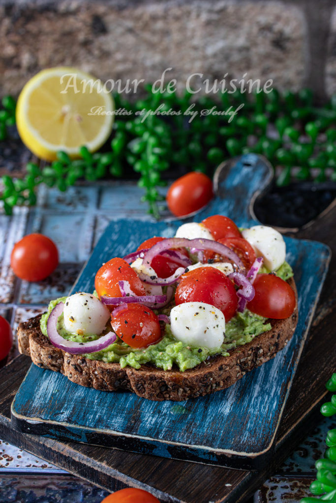Whole grain avocado toast topped with cherry tomatoes, mini mozzarella balls, and sliced red onions, served on a rustic blue wooden board with a background of lemon, cherry tomatoes, and greenery