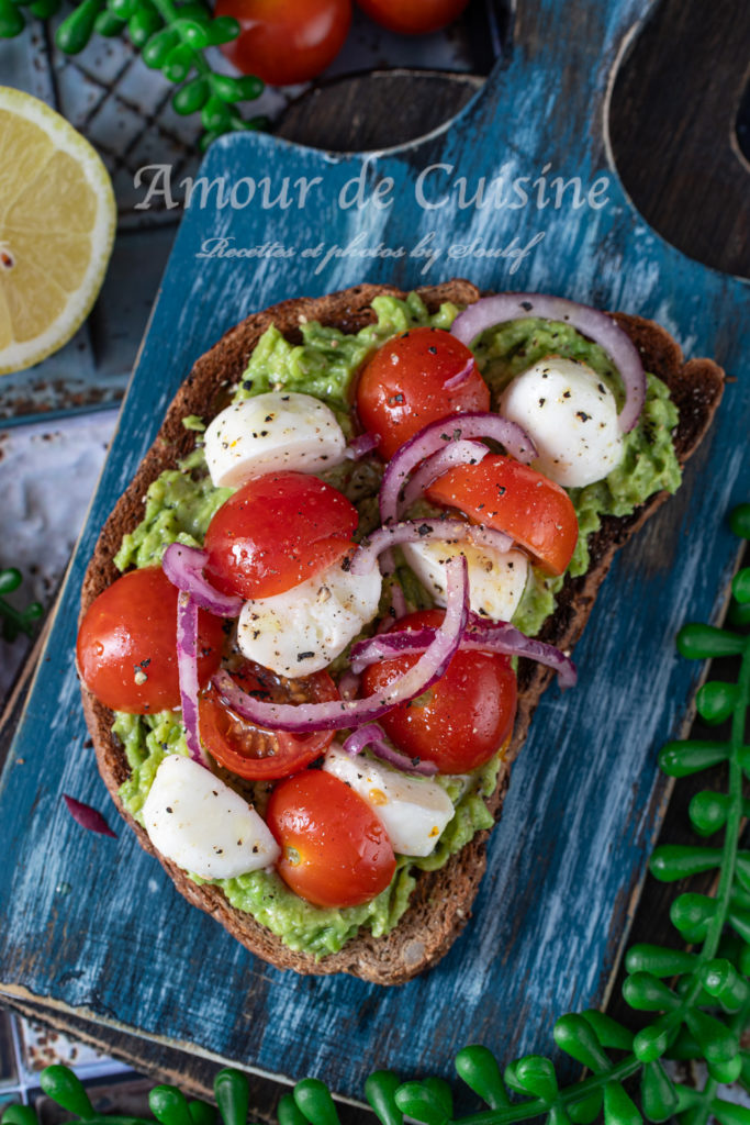 Caprese-style avocado toast topped with halved cherry tomatoes, mini mozzarella balls, and thin slices of red onion on whole grain bread, served on a rustic blue wooden board with lemon and tomatoes in the background