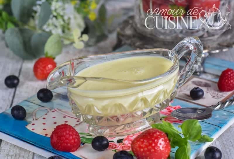 A glass sauce boat filled with creamy homemade vanilla custard (crème anglaise), placed on a decorative napkin surrounded by fresh berries including raspberries, strawberries, and blueberries. A spoon rests nearby, and a floral arrangement in the background adds a fresh, inviting touch