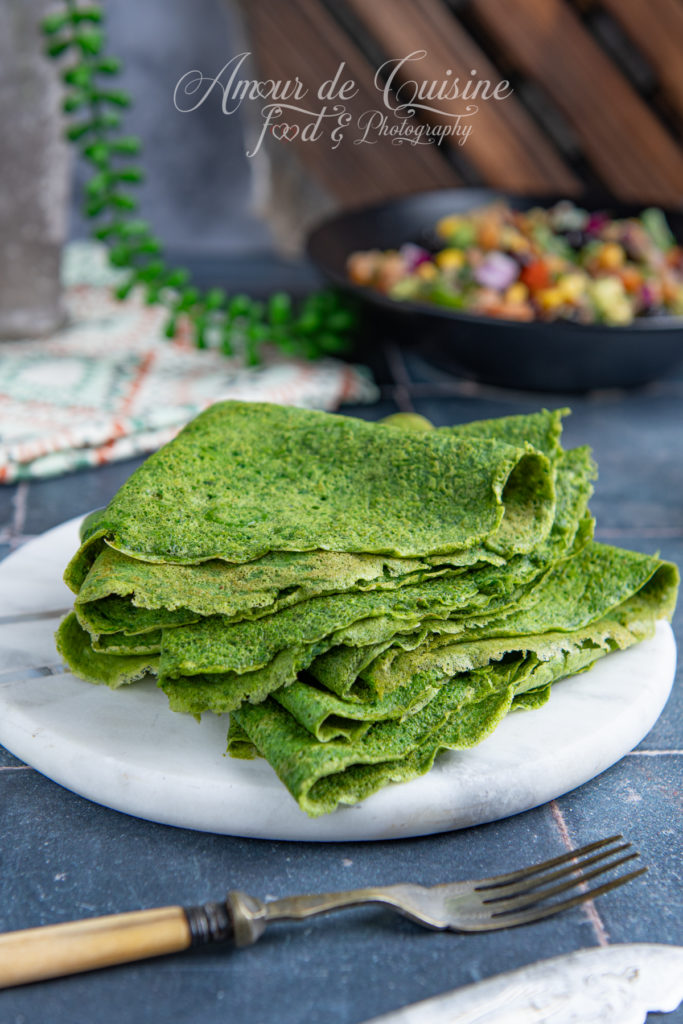 A stack of folded homemade spinach tortillas with a vibrant green color, neatly arranged on a round marble plate, with a rustic fork in the foreground and a colorful vegetable salad blurred in the background