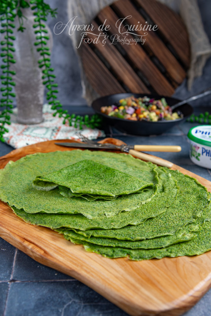 A stack of vibrant green spinach tortillas neatly arranged on a wooden cutting board, with a knife and fork beside them; a bowl of colorful vegetable salad and a tub of cream cheese visible in the background