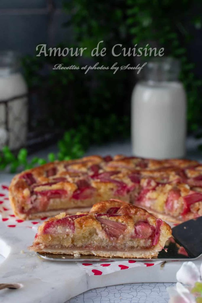 A close-up of a slice of rhubarb and almond cream tart on a white serving board with red hearts. The tart has a golden crust, a layer of almond filling, and vibrant pink rhubarb pieces on top. A bottle of milk and green foliage are blurred in the background, creating a cozy and inviting atmosphere