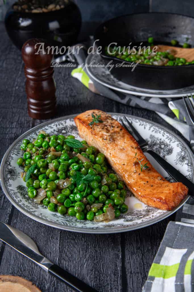 Air-fried salmon fillet served with sautéed green peas and caramelized onions, garnished with fresh mint, on a decorative plate with a fork and knife beside it. A skillet with more peas and a wooden spoon is visible in the background