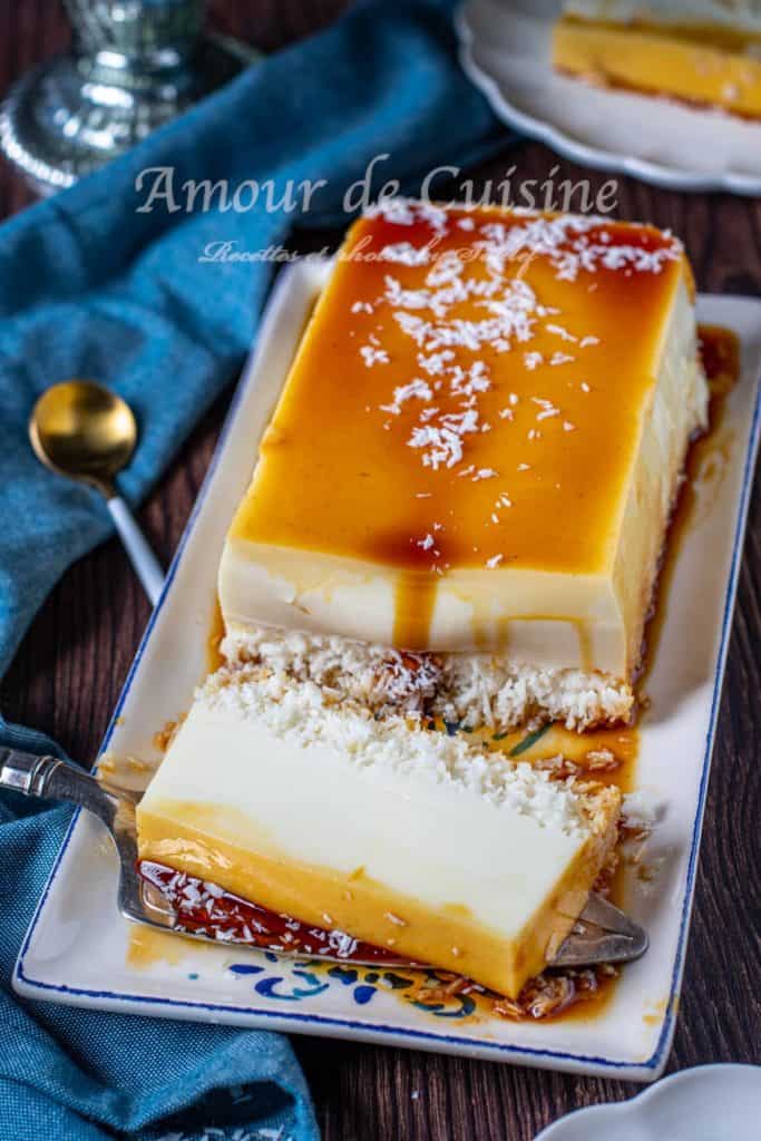 A rectangular Caribbean or french entilles coconut flan (Flan Coco Antillais) topped with caramel sauce and shredded coconut, partially sliced to reveal its creamy layers, served on a decorative rectangular plate with a gold spoon and blue napkin nearby.