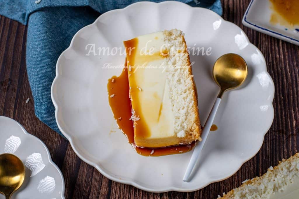 A slice of Caribbean coconut flan (Flan Coco Antillais) served on a scalloped white plate, showcasing its creamy custard layer, caramel topping, and shredded coconut base, with a gold and white spoon placed beside it