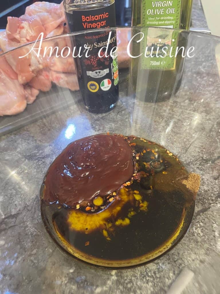 A glass bowl filled with ingredients for a barbecue chicken marinade, including barbecue sauce, balsamic vinegar, olive oil, crushed red pepper flakes, and spices, with raw chicken wings and condiment bottles visible in the background