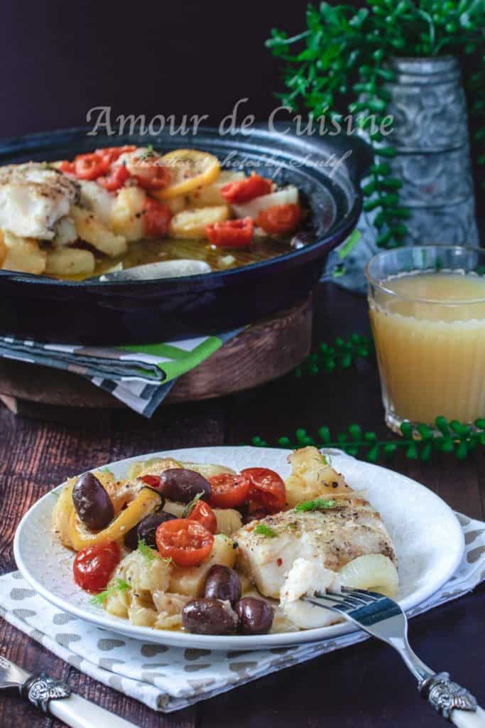 A beautifully presented plate of fennel, olive, and smoked haddock tagine, served with roasted cherry tomatoes and tender potato slices. The dish is garnished with fresh herbs and accompanied by a glass of juice. In the background, a traditional black tagine pot filled with the same colorful and aromatic meal sits on a wooden surface, with green decorative plants adding a cozy touch