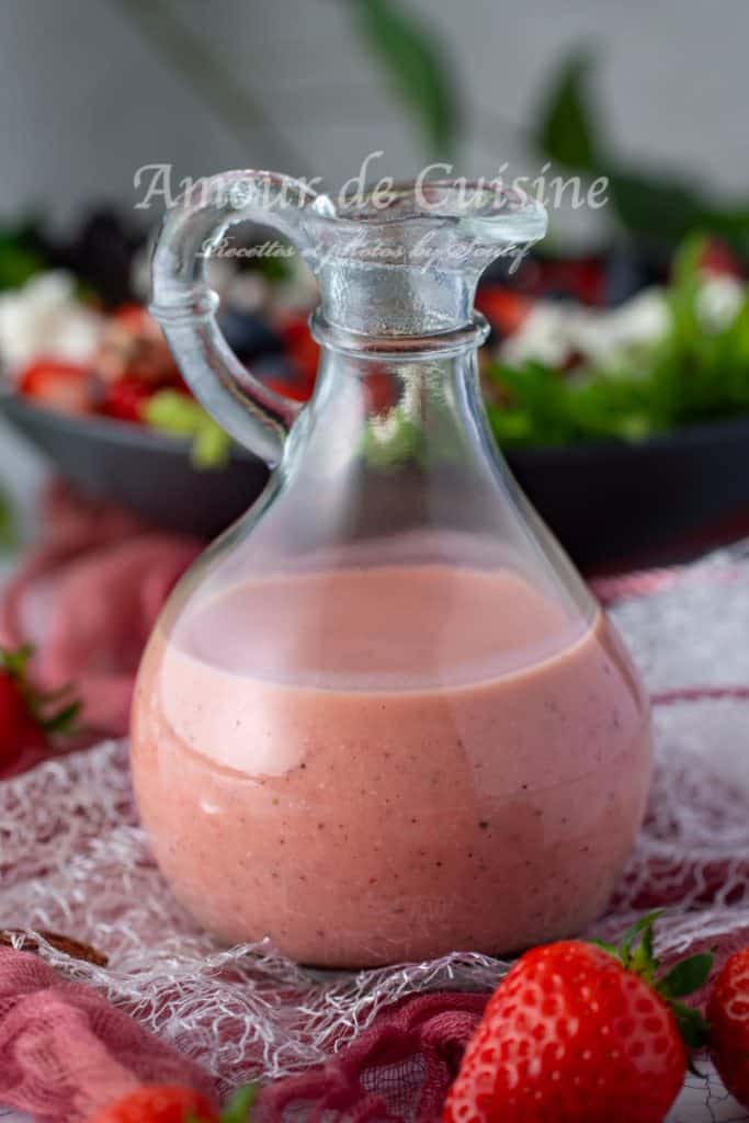 A glass bottle filled with creamy pink strawberry vinaigrette, placed on a textured white and red cloth with fresh strawberries scattered around. In the blurred background, a vibrant strawberry and feta salad is visible, adding a fresh and appetizing touch to the scene