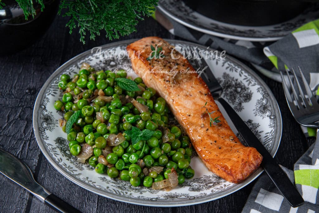 A beautifully plated dish of pan-seared salmon fillet served with buttery sautéed green peas and caramelized shallots, garnished with fresh mint, on a decorative floral plate set on a rustic black wooden table.