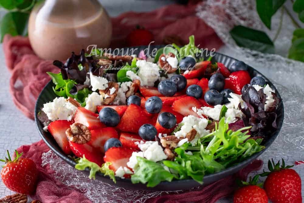 A colorful summer salad served in a black bowl, featuring fresh mixed greens, sliced strawberries, whole blueberries, crumbled feta cheese, and chopped pecans. The salad is arranged beautifully, with a bottle of creamy strawberry vinaigrette blurred in the background and fresh strawberries scattered around the scene