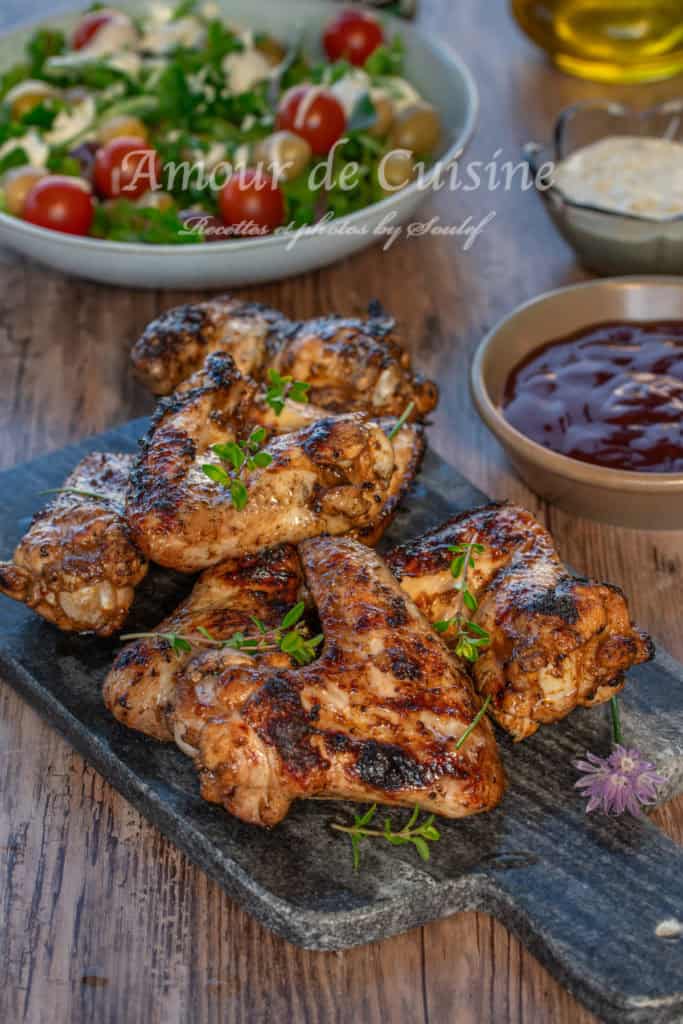 Grilled barbecue chicken wings served on a slate board, garnished with fresh herbs, accompanied by a bowl of barbecue sauce, creamy dip, and a fresh salad with cherry tomatoes and green olives in the background