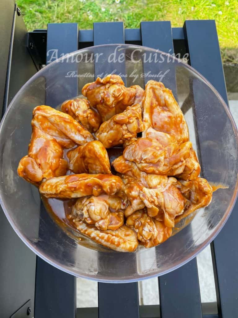A glass bowl filled with raw chicken wings thoroughly coated in a rich, glossy barbecue marinade, sitting on a black outdoor table with green grass visible in the background