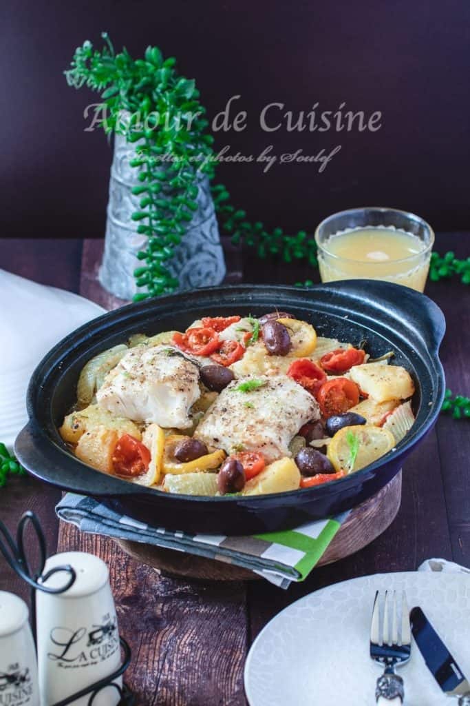 A hearty fennel, olive, and haddock tagine served in a traditional black tagine dish. The dish features golden potato slices, juicy cherry tomatoes, black olives, and two flaky haddock fillets, all gently cooked in a fragrant broth. The scene is styled with rustic charm—wooden table, green napkins, and a glass of juice, with fresh greenery in the background adding a cozy, homemade feel. An empty plate and cutlery are ready for serving.
