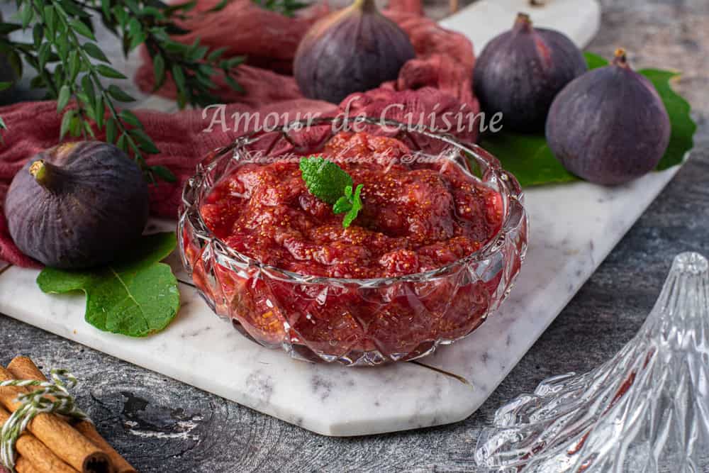 Un bol en verre taillé rempli de compote de figues sans sucre, à la texture épaisse et brillante, décoré d’une feuille de menthe fraîche. Le bol repose sur une planche de marbre blanc entourée de figues fraîches entières, de feuilles de figuier, de bâtons de cannelle et d’un tissu rouge. Le couvercle du bol est posé à côté, et l’arrière-plan présente un décor rustique et naturel