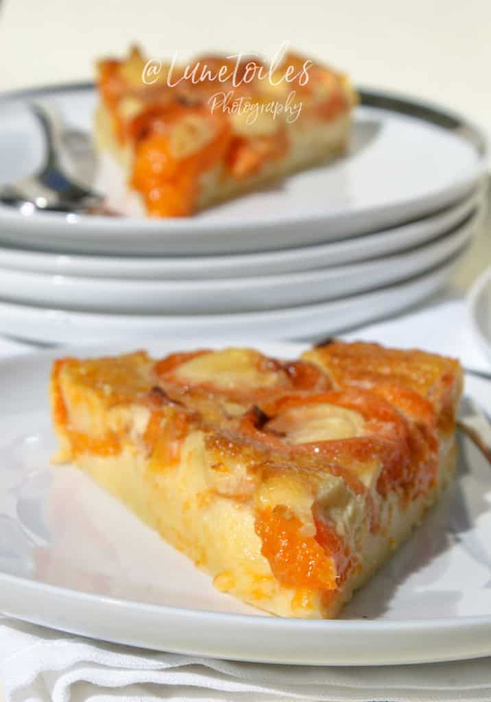 A close-up of a slice of apricot clafoutis served on a white plate, showcasing its golden-brown top and vibrant orange apricot halves embedded in a smooth, custard-like base. The plate rests on a white napkin, with another slice in soft focus stacked on a pile of plates in the background. The image is bright and sunlit, with the watermark "@Lunetoiles Photography" centered at the top