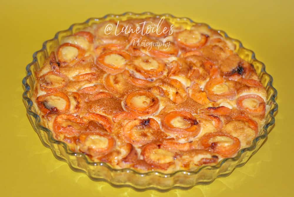 A freshly baked apricot clafoutis in a clear, fluted glass baking dish, set against a vibrant yellow background. The dessert features halved apricots nestled in a golden, custard-like batter with lightly browned edges and a glossy finish. The clafoutis appears rich and inviting, with the apricots slightly caramelized on top. The watermark "@Lunetoiles Photography" is visible across the center