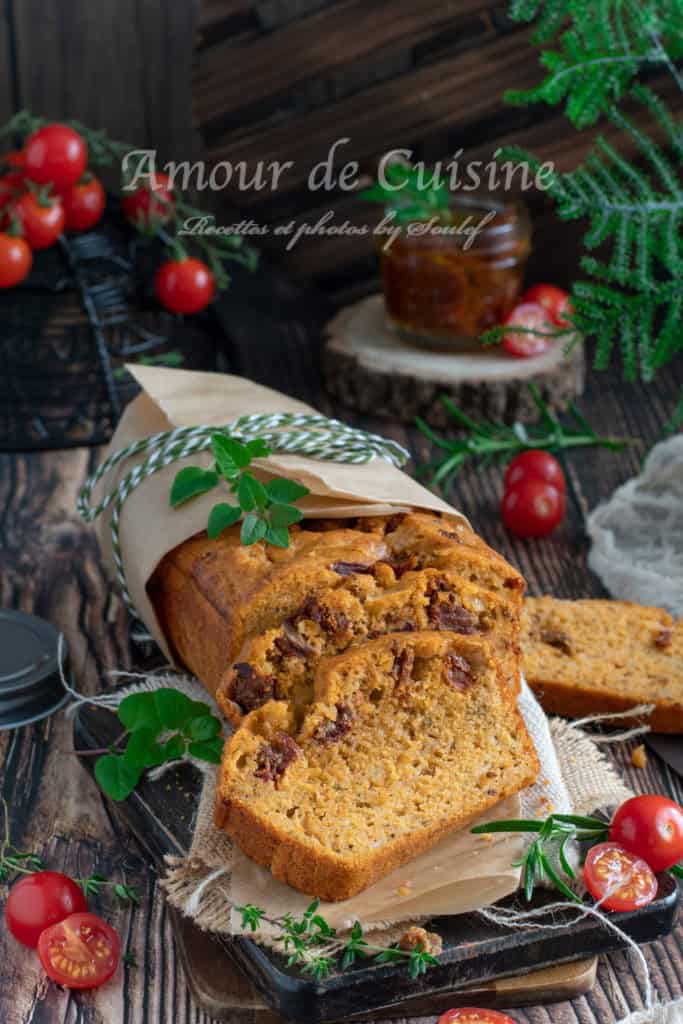Sliced savoury cake with pesto rosso and sun-dried tomatoes, wrapped in parchment and string, presented on a rustic wooden board with cherry tomatoes, herbs, and a jar of pesto in the background