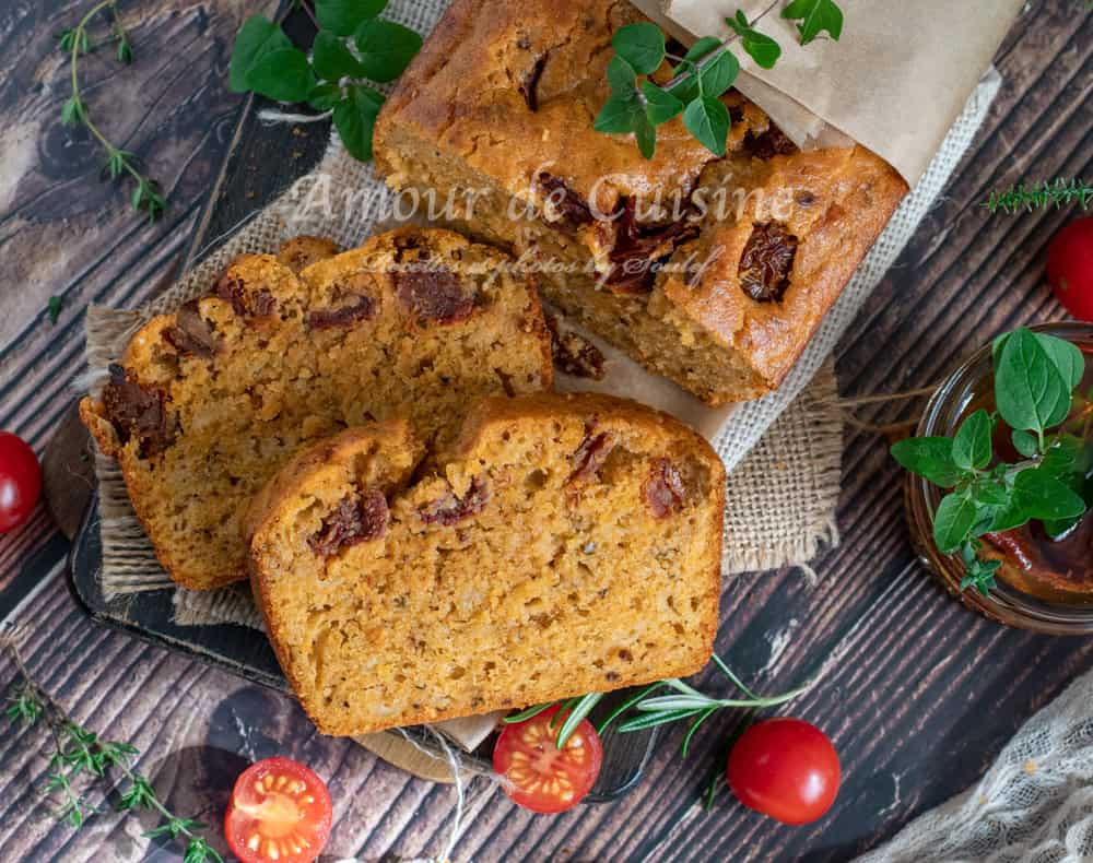 Top view of sliced savoury cake with pesto rosso and sun-dried tomatoes on a rustic wooden surface, surrounded by fresh herbs, cherry tomatoes, and a jar of pesto