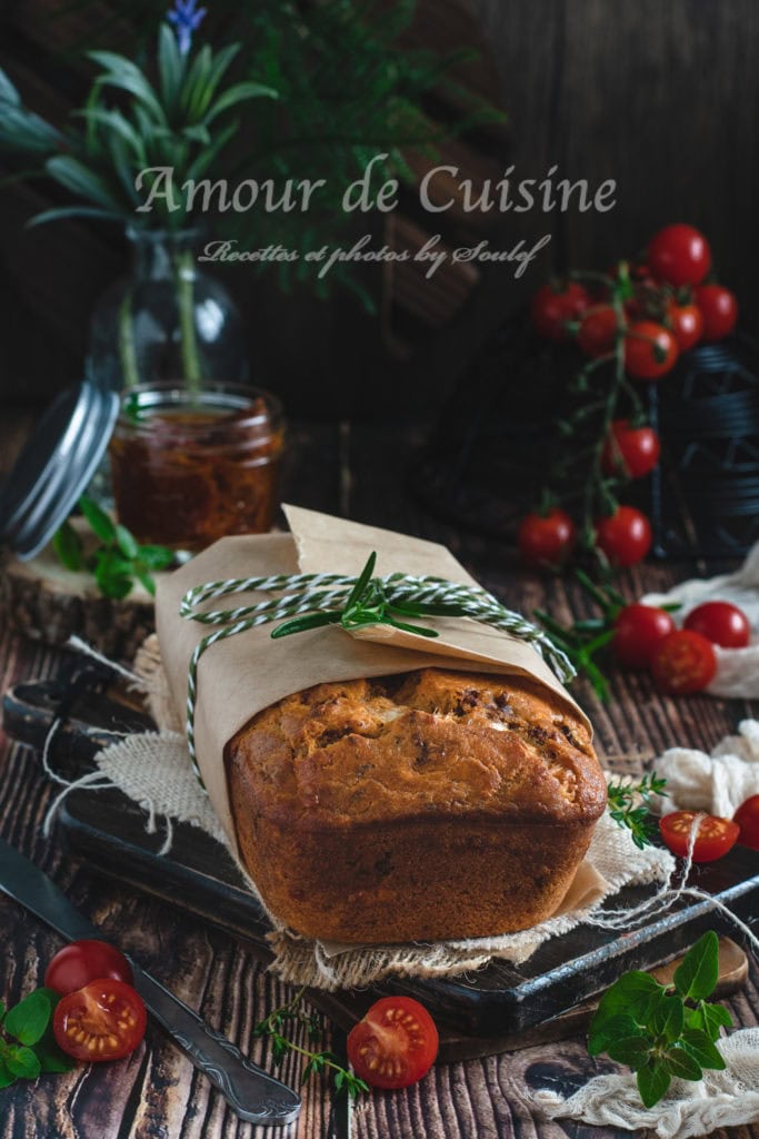Whole savoury cake with pesto rosso and sun-dried tomatoes, wrapped in parchment paper and tied with string, displayed on a rustic wooden board surrounded by cherry tomatoes, herbs, and a jar of pesto