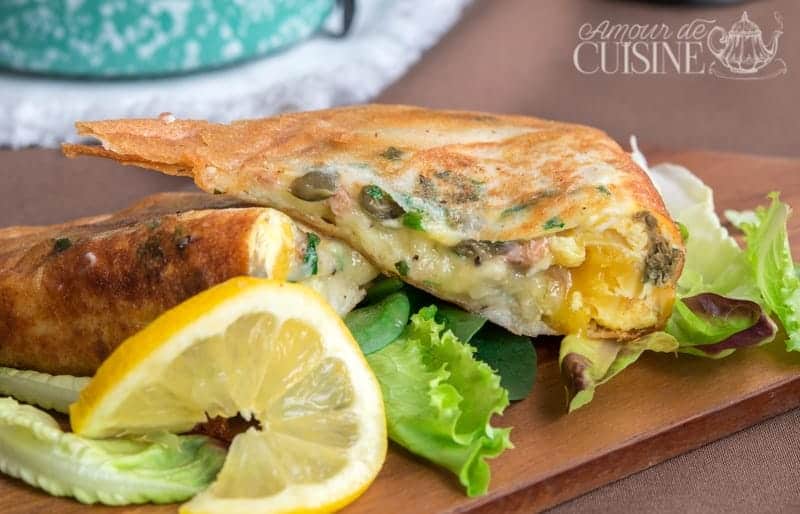 Golden Tunisian brik cut open to reveal a soft egg yolk, tuna, capers, and herbs, served with mixed greens and lemon wedge on a wooden board