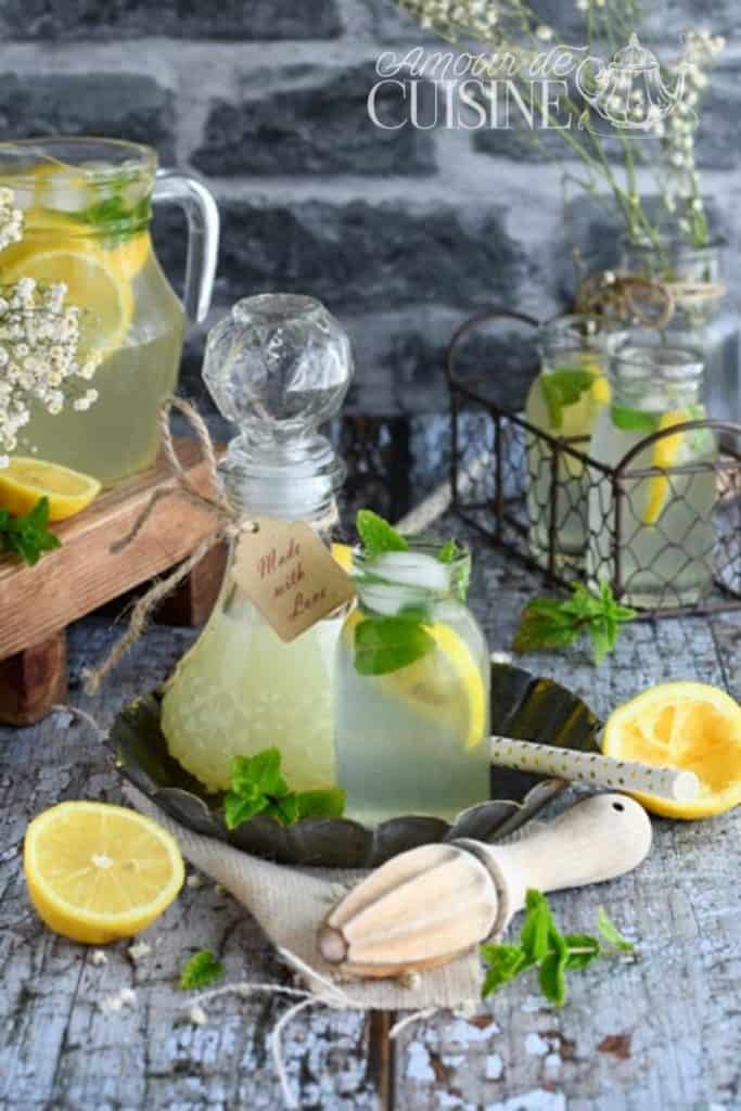 A rustic summer setup featuring homemade lemonade in a glass pitcher with lemon slices and mint, surrounded by vintage-style glass bottles, fresh lemon halves, mint sprigs, a wooden citrus juicer, and delicate white flowers, all arranged on a weathered wooden table against a gray brick background