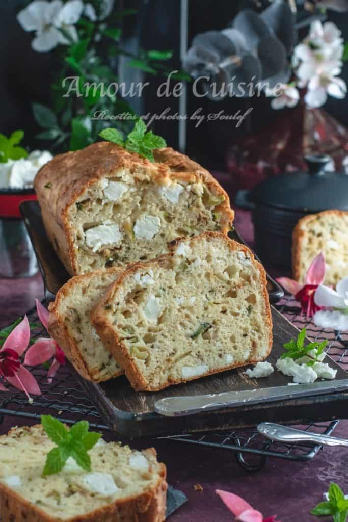Front view of a sliced zucchini and feta savoury cake on a wooden board, showing its golden crust and fluffy interior, garnished with fresh mint and surrounded by scattered feta and flowers