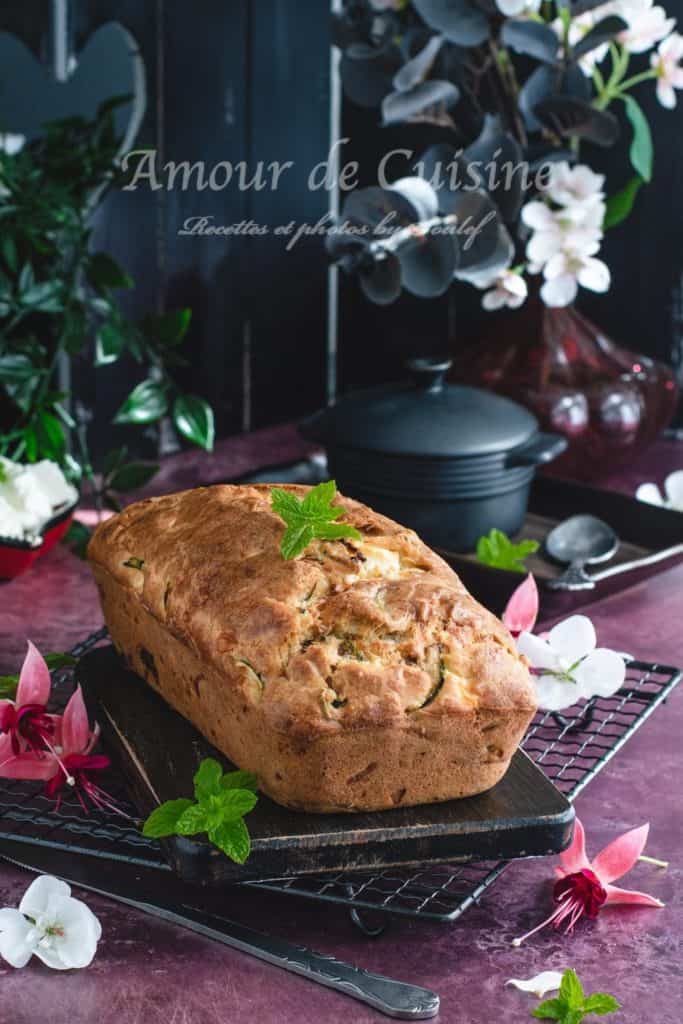 Whole zucchini and feta savoury loaf cake displayed on a wooden board, golden brown with a mint garnish on top, surrounded by flowers and kitchenware in a rustic setting.